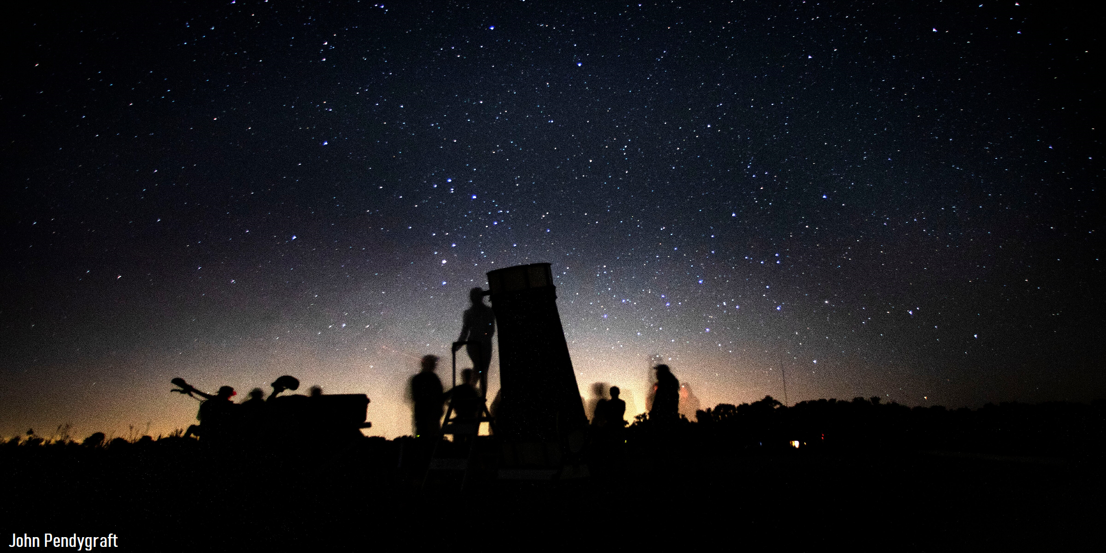 A silhouette of a large telescope and several people against a starry sky with the glow of the horizon behind them